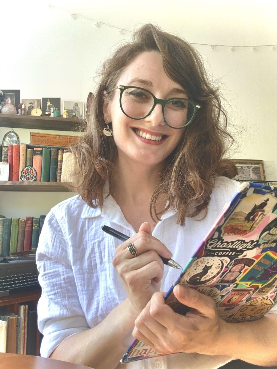 Ashley smiling in green round glasses and a white linen shirt, pen in hand, holding a sticker-covered journal in front of her bookshelves.