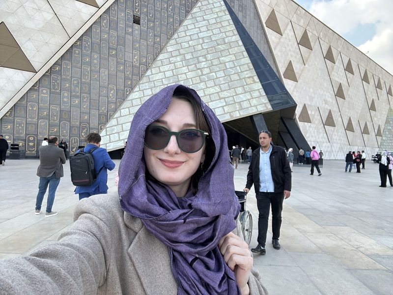 Ashley in a purple headscarf and sunglasses taking a selfie in front of a grand museum facade.