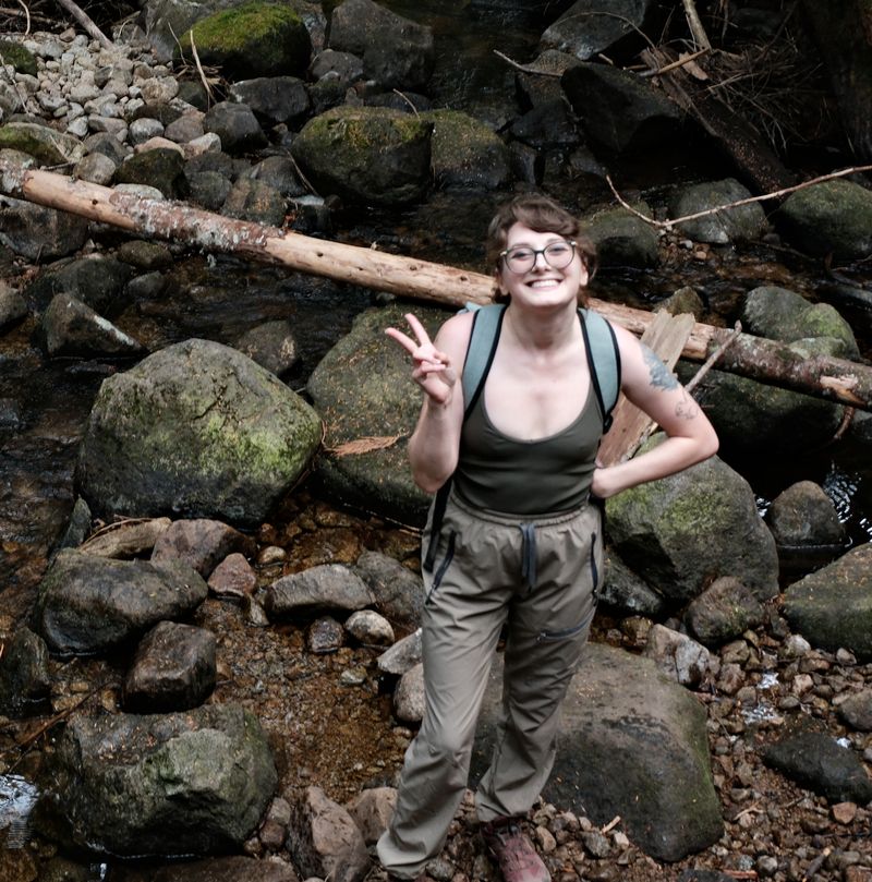 Ashley in hiking gear, grinning and making a peace sign among mossy creek rocks.