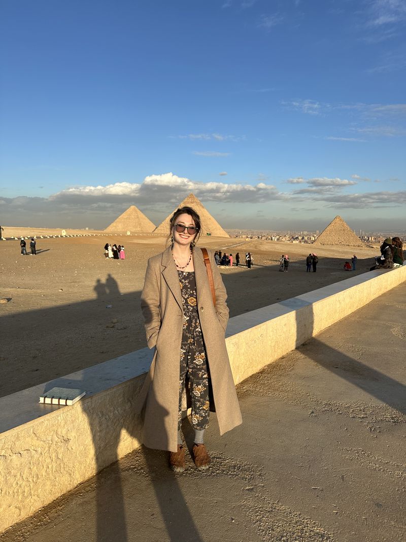 Ashley in a long coat standing on a terrace, with the pyramids of Giza behind her at sunset.
