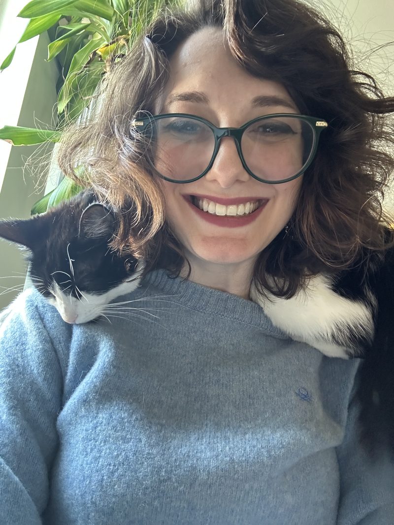 Ashley smiling in a blue sweater, with Rette, a black and white cat, draped across her shoulders.