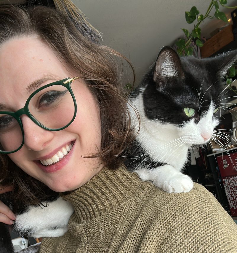 Ashley smiling in an olive sweater, with Rette, a black and white cat, perched on her shoulder.